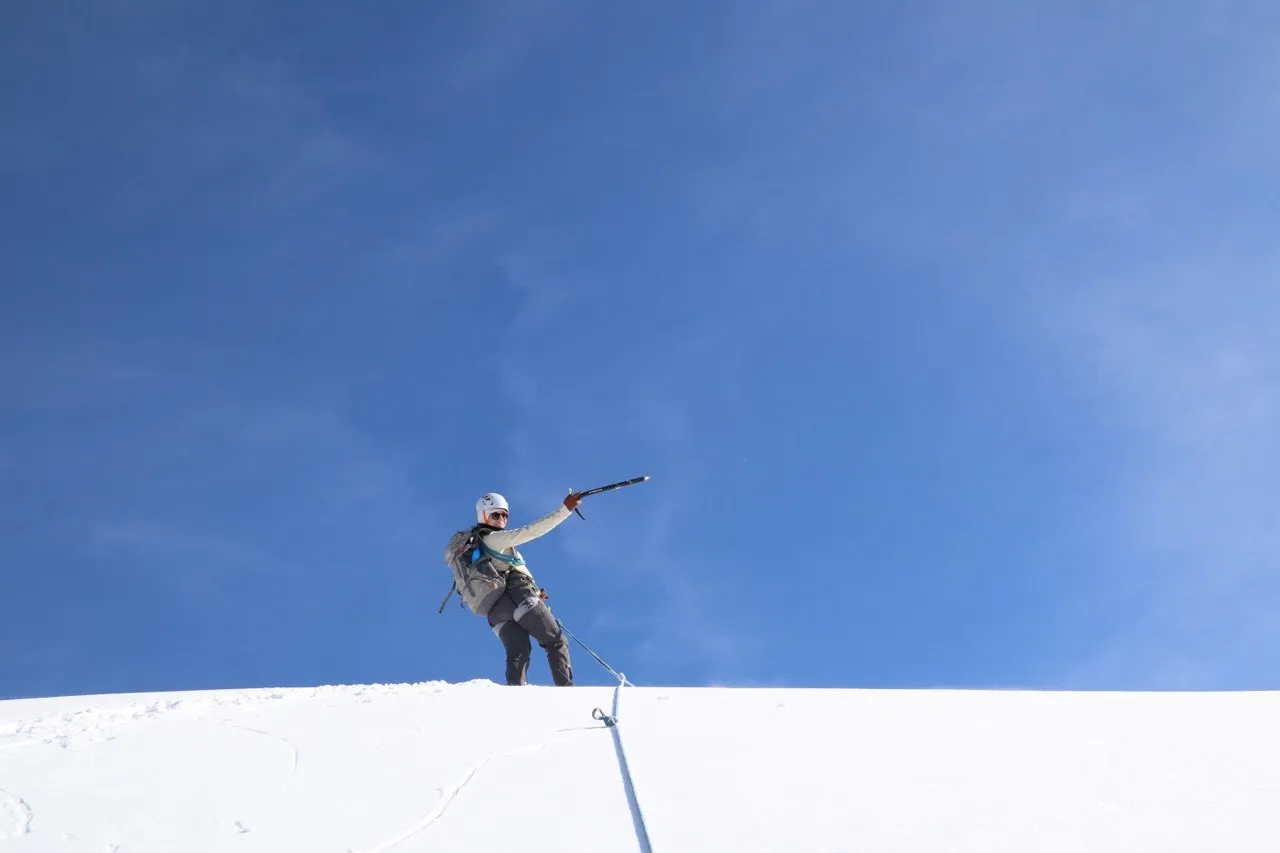 Hiker on the snowy summit dome raising an ice axe against a blue sky.