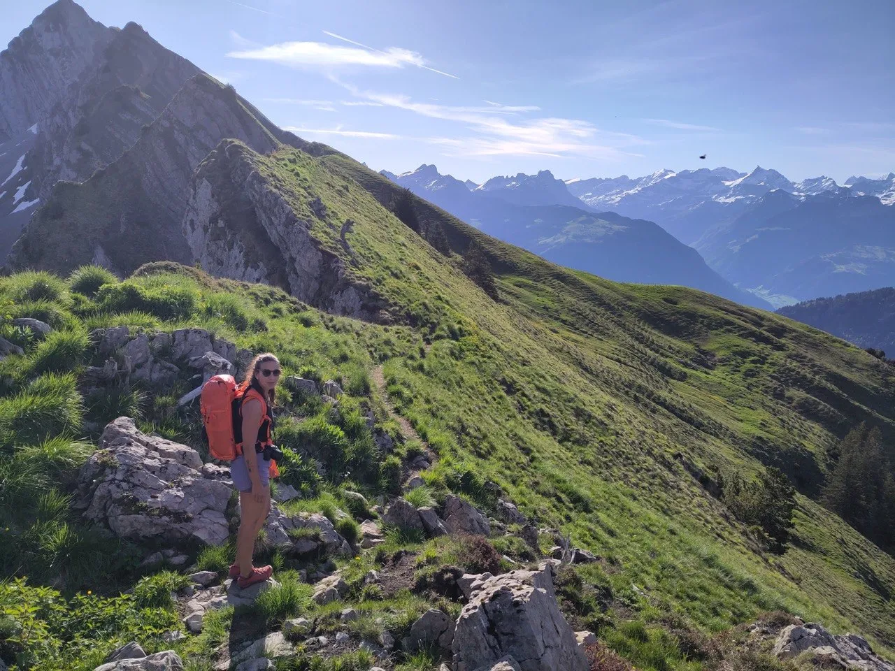 A hiker with an orange backpack pauses on a grassy ridge, mountains rolling out behind her.