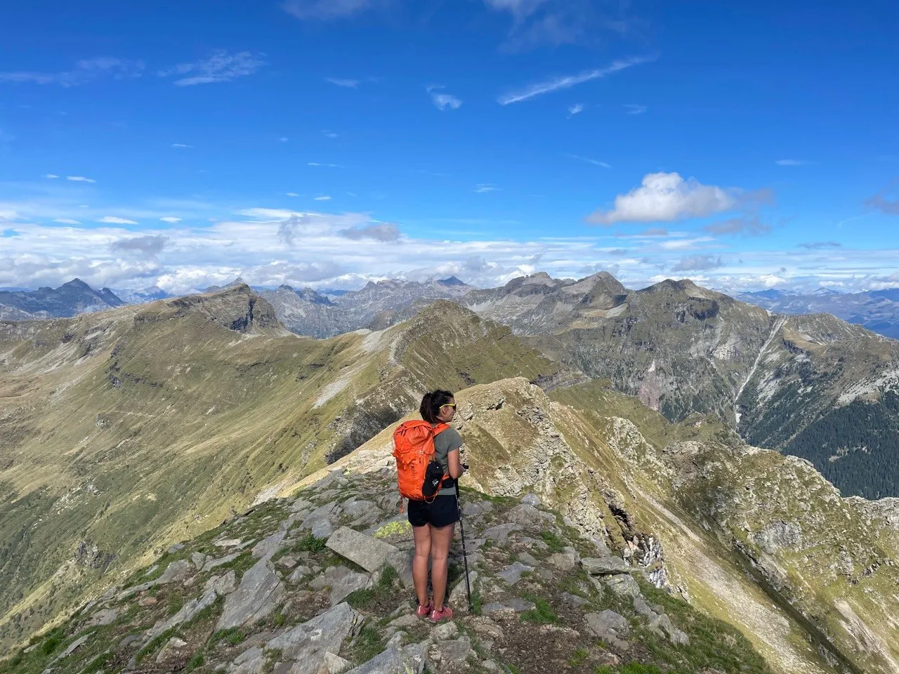 Hiker with an orange backpack looking out over a sea of green-and-grey ridges from a rocky crest.