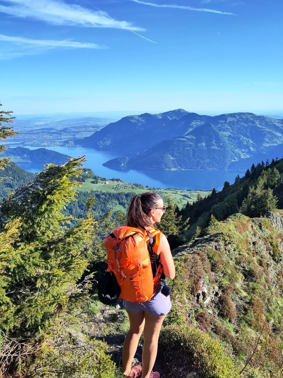 A hiker looks out over a lake framed by mountains from a high ridge, orange backpack and all.