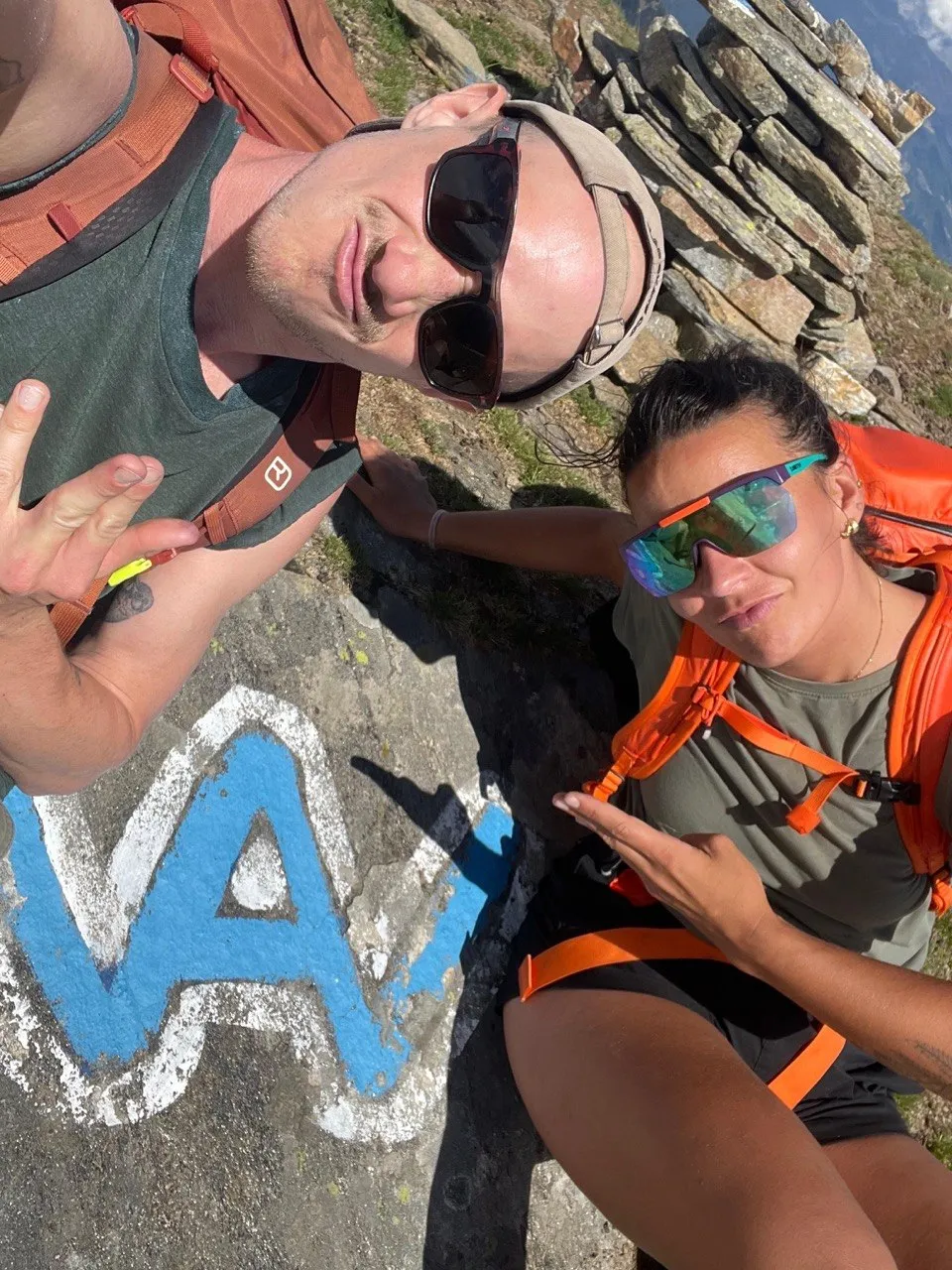 Two hikers taking a selfie beside a painted blue Via Alta trail marker on the rock.