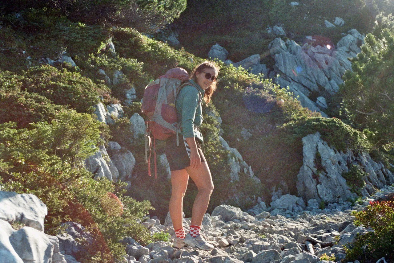 A hiker with a large backpack and sunglasses stands smiling in warm morning light among mountain pines and pale rock.