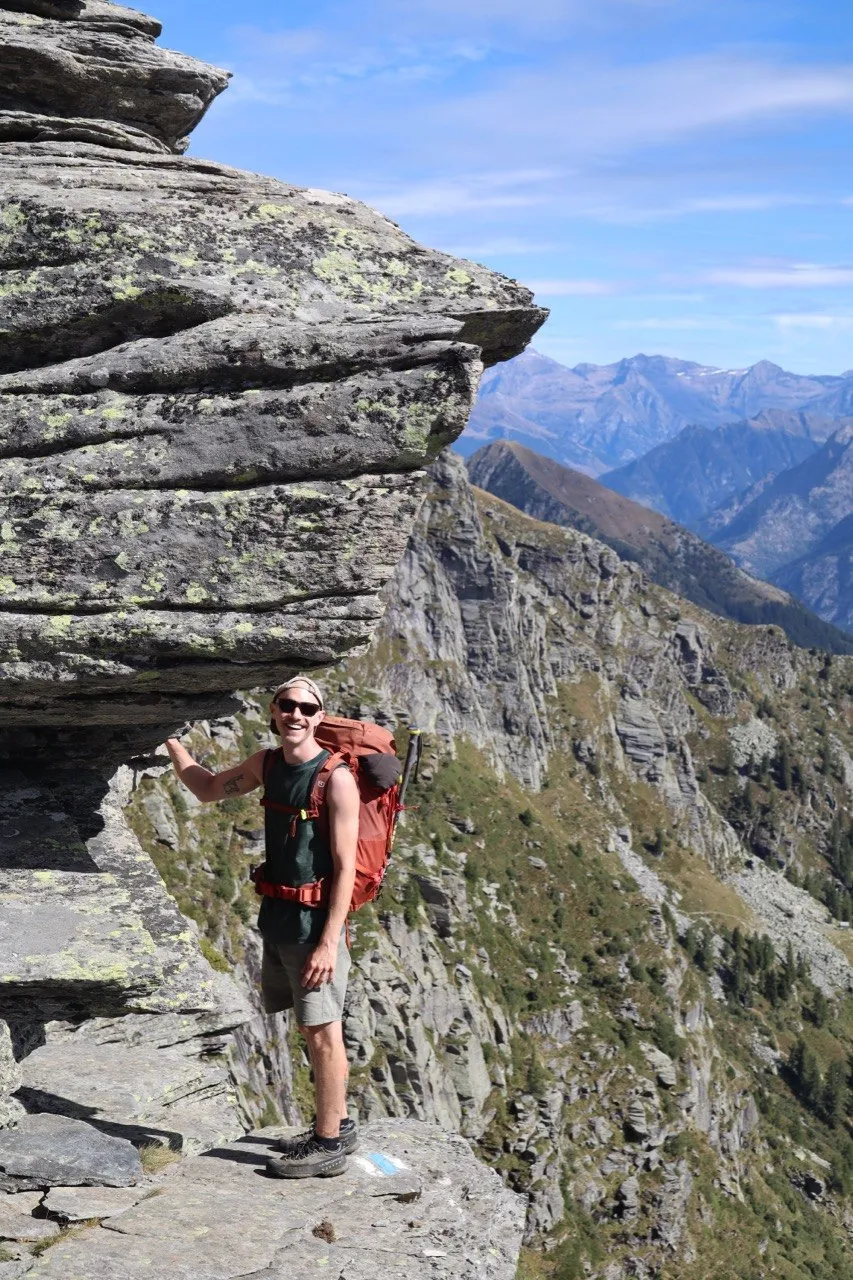 Hiker with a red backpack posing under a large layered rock overhang, rugged peaks behind.