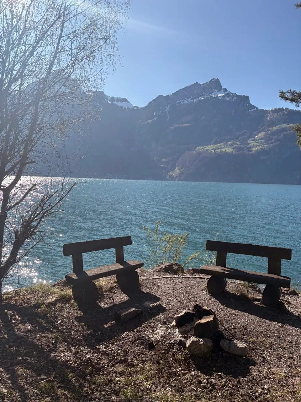 Two wooden benches and a fire pit on a gravel lakeshore, with the turquoise lake and snow-capped mountains behind — the grill spot before Tellskapelle.