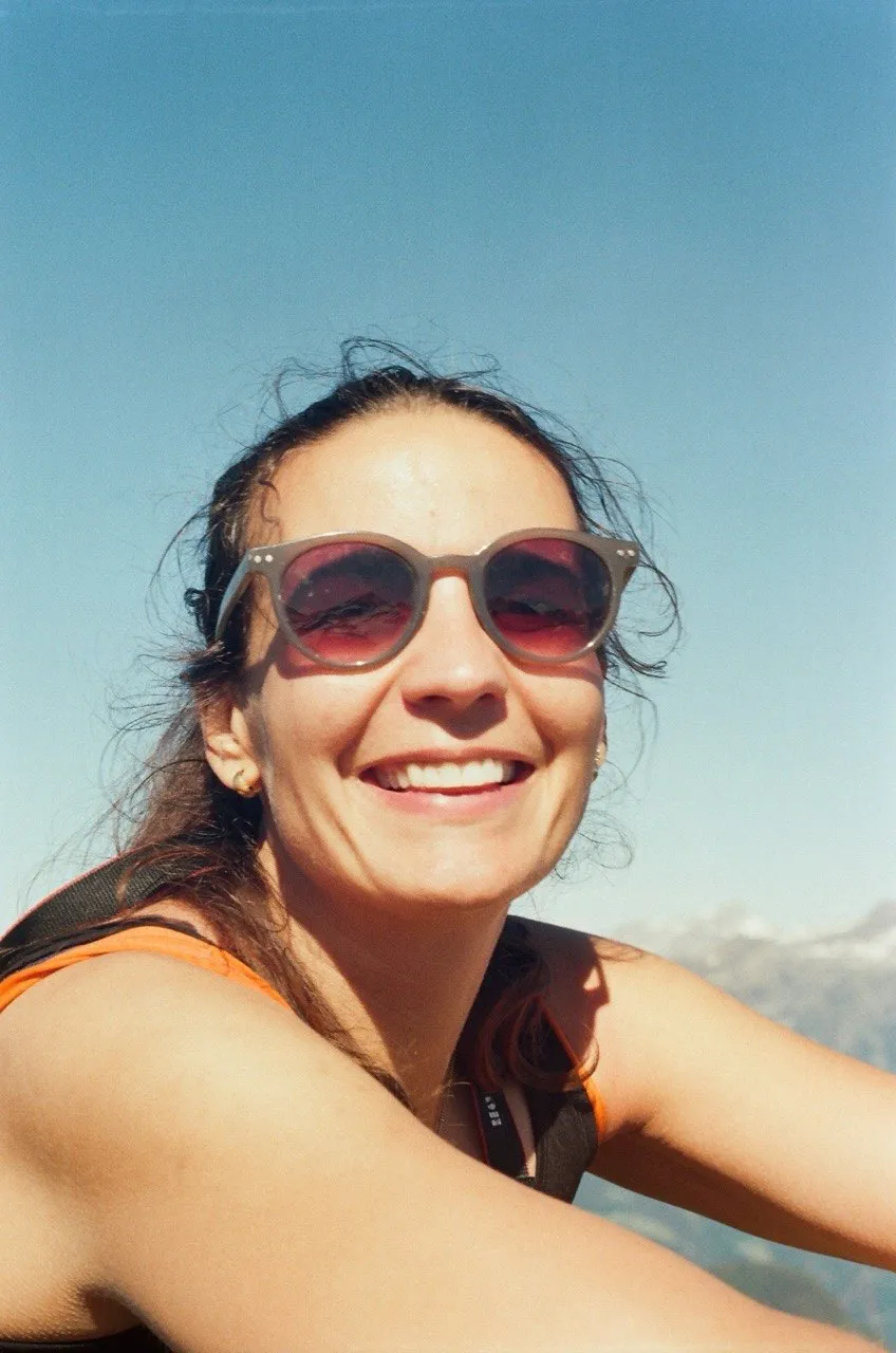 A smiling hiker in sunglasses on the summit, with distant snow-capped peaks under a deep blue sky.