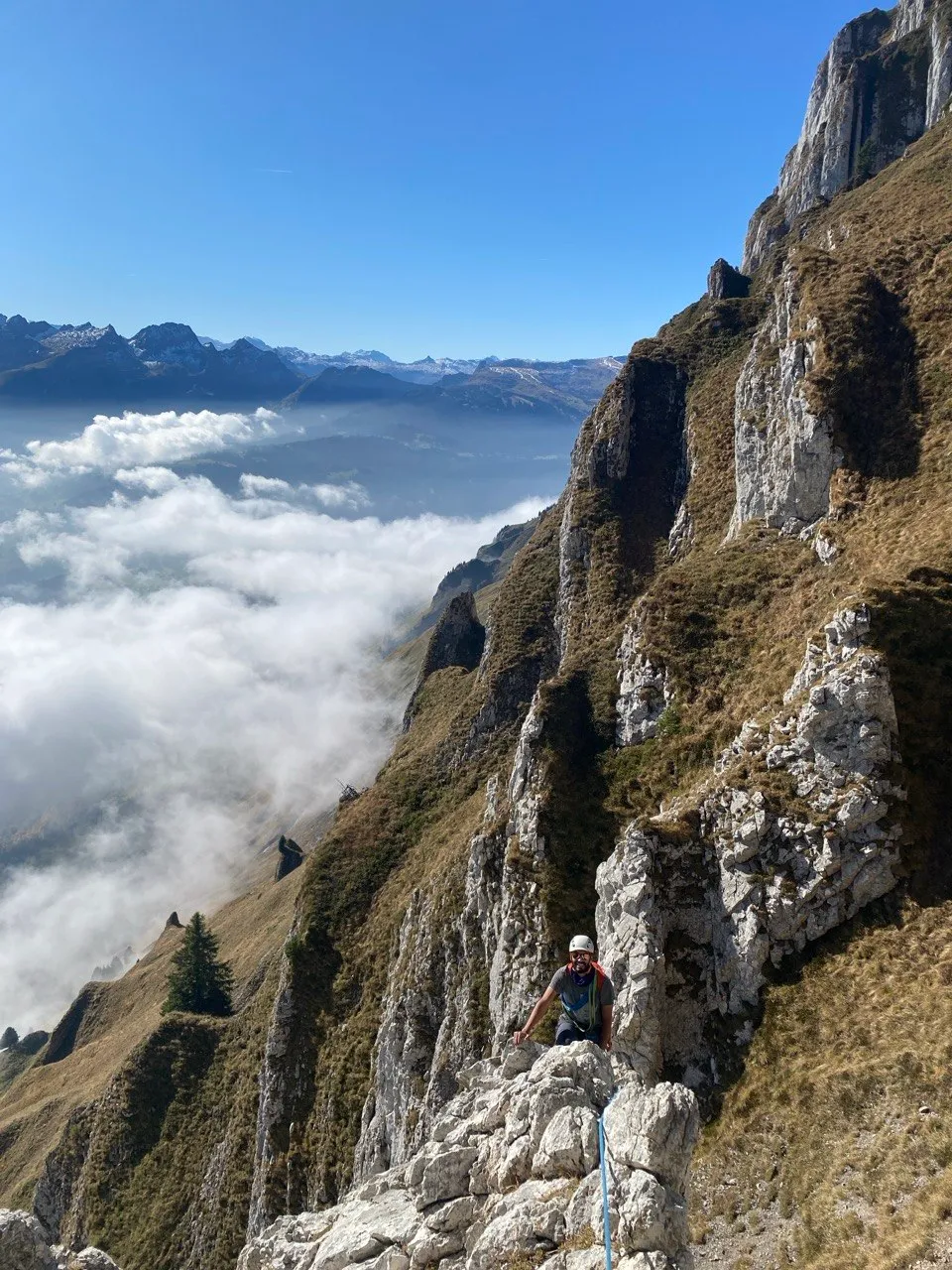 A helmeted hiker on a narrow white limestone ledge, a deep sea of fog and distant ridges far below.