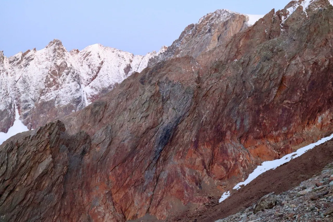 Close-up of a red-and-grey rock wall with snowy peaks behind in the dawn light.