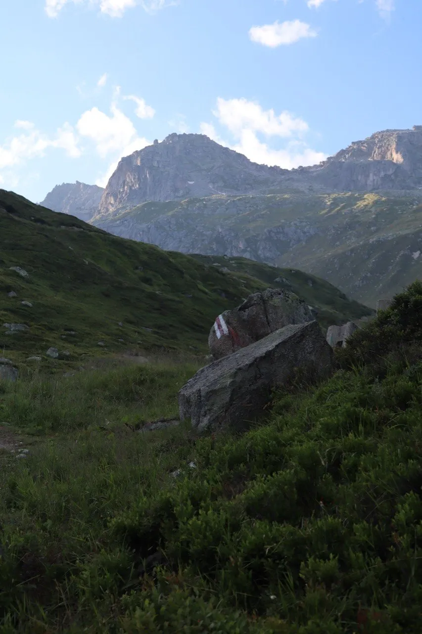 A red-and-white trail marker painted on a boulder beside a grassy path, with rugged peaks rising under a blue sky.