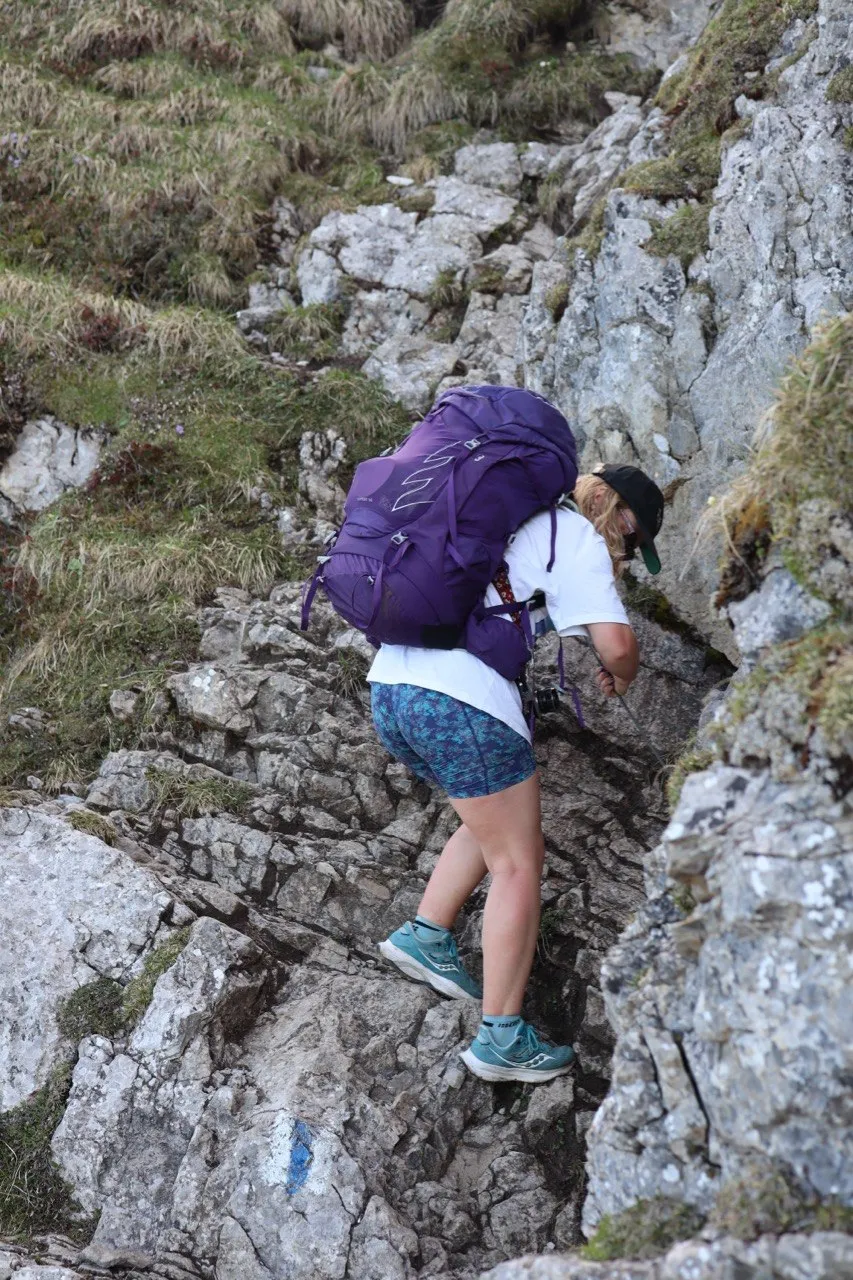 A hiker in a purple backpack scrambles up a narrow rocky gully, hands and feet on the rock.