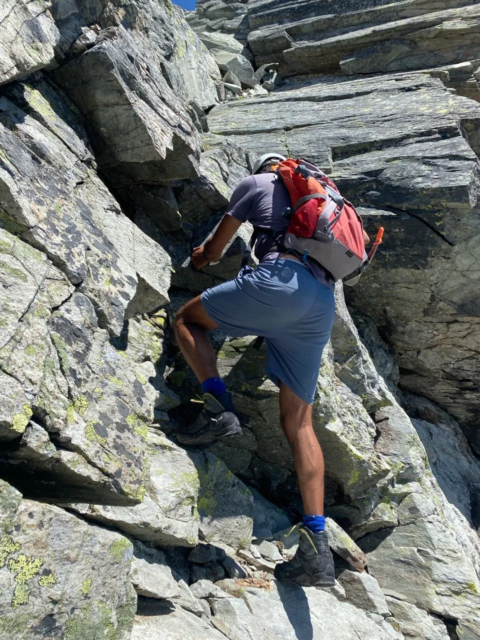 A hiker with an orange backpack scrambles up a steep, slabby rock step.