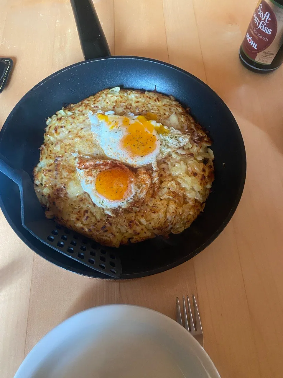 Overhead view of a pan with rösti and two fried eggs, a fork and plate ready on a wooden table.