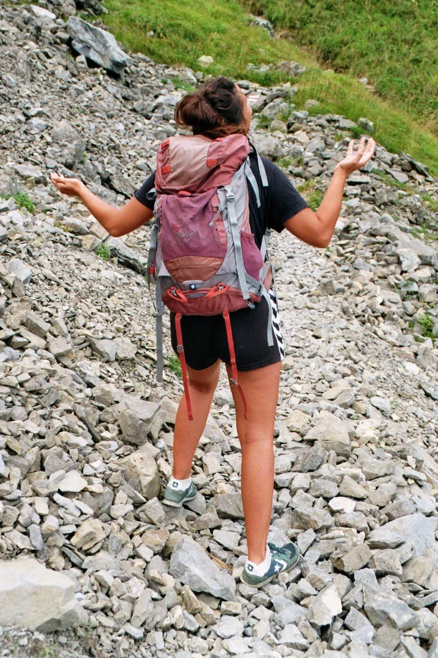A hiker with a red backpack balancing arms-out across a field of loose scree.