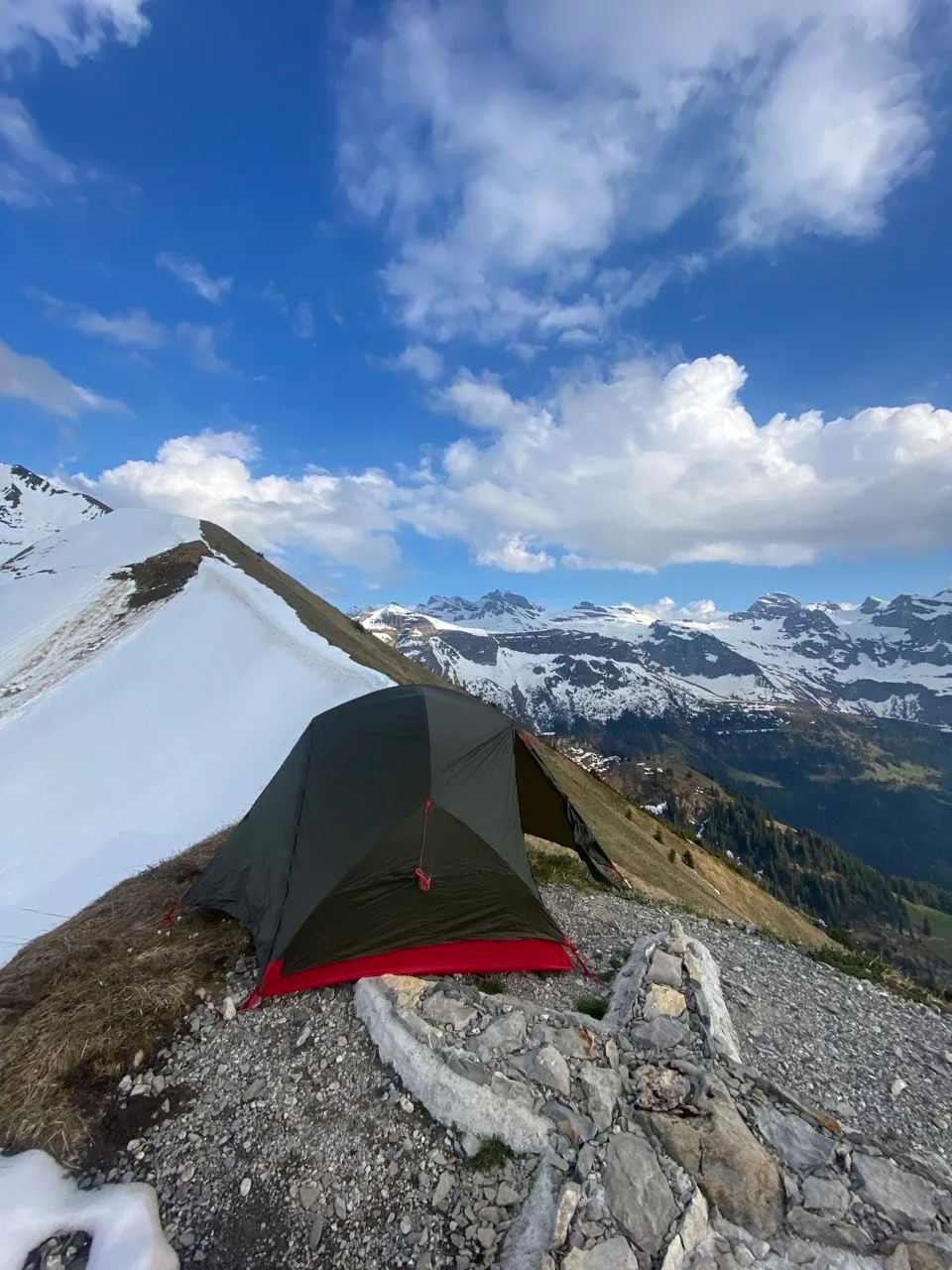 A green tent pitched on a narrow ridge with patches of snow, backed by a wide panorama of snow-covered peaks under a blue sky.