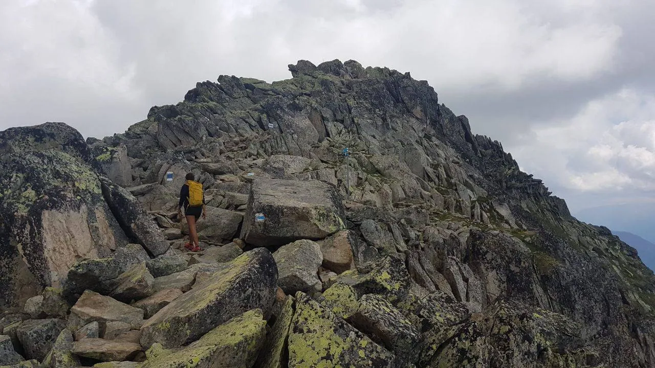 A hiker with a yellow backpack climbs over boulders and scree toward a jagged rock ridge, blue-and-white trail blazes on the blocks.