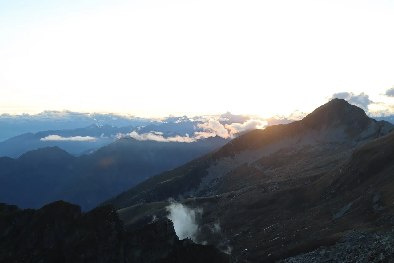 Sunrise over layered mountain ridges, the low sun breaking behind a dark peak with clouds drifting through the valleys below.