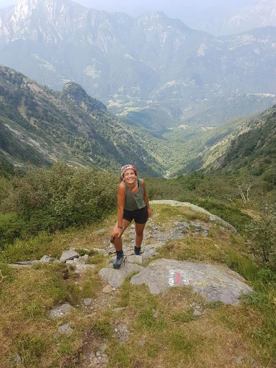 A hiker in a headscarf stands smiling on a rocky knoll with a white-and-red waymark, a deep forested valley and hazy southern-Alpine peaks behind.