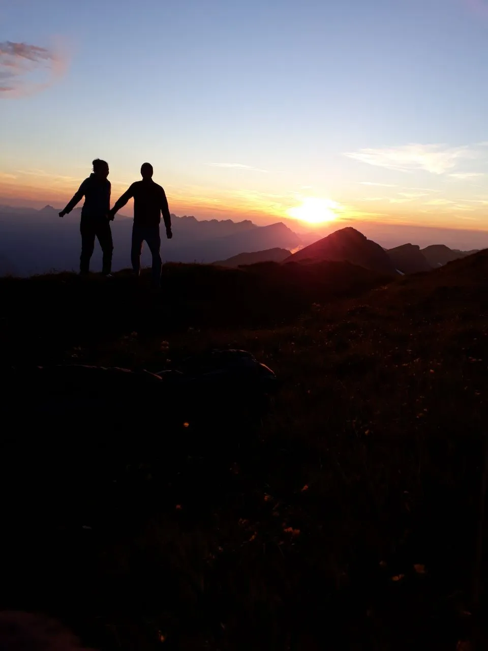 Two hikers silhouetted on the summit ridge against an orange sunset over a sea of peaks.