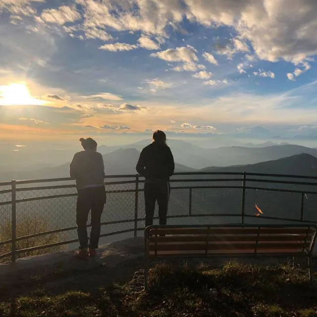 Dos personas de pie junto a la barandilla de un mirador contemplan cadenas montañosas brumosas hacia un sol bajo, con un banco en primer plano.