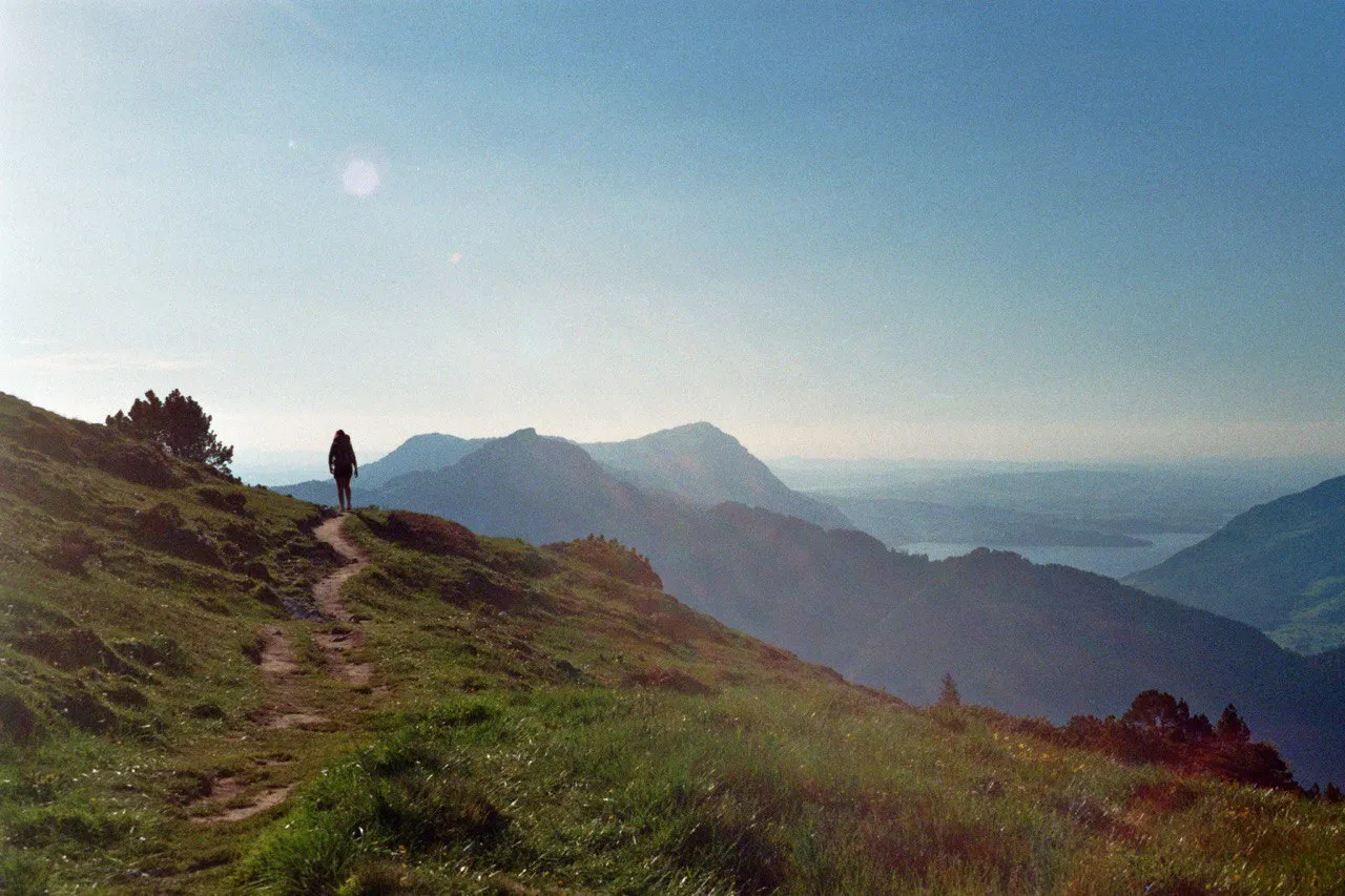 A hiker as a small silhouette on a narrow grassy path, layered ridges and the hazy, glinting lake spread out ahead in the morning backlight.