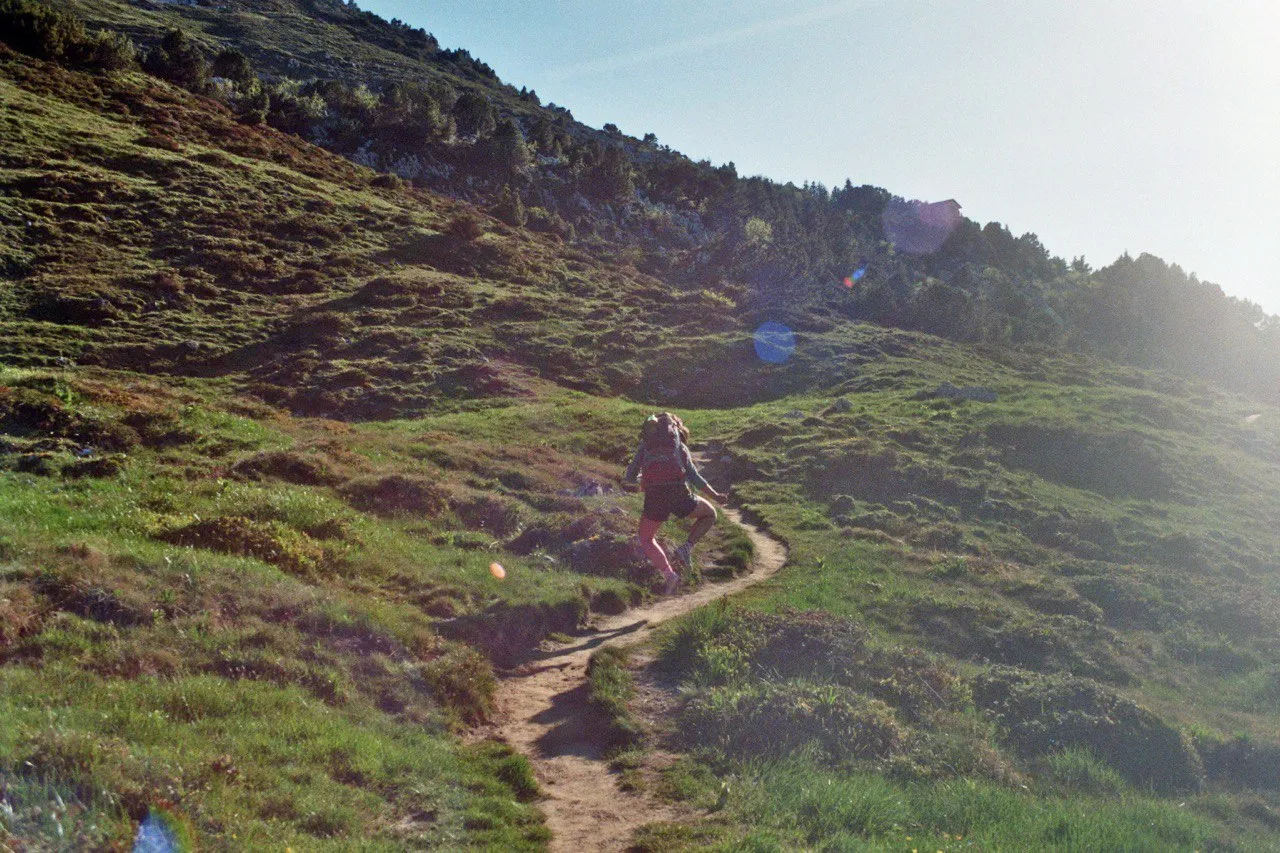 A hiker runs down a winding path through a sunlit green alp, lens flares scattered across the backlight.