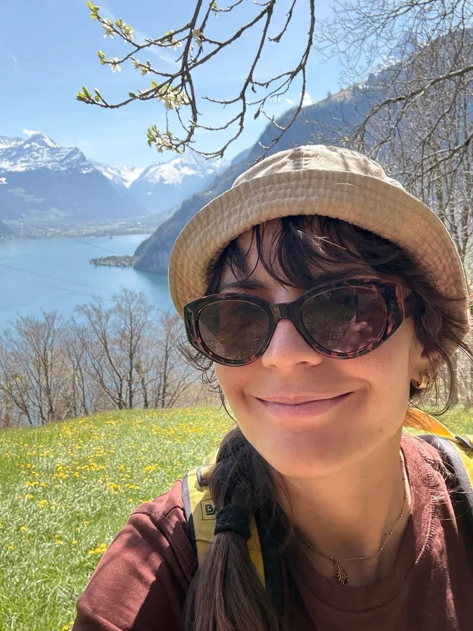 Selfie of the hiker in sunglasses and a bucket hat on a spring meadow full of dandelions, the lake and snowy peaks behind her.