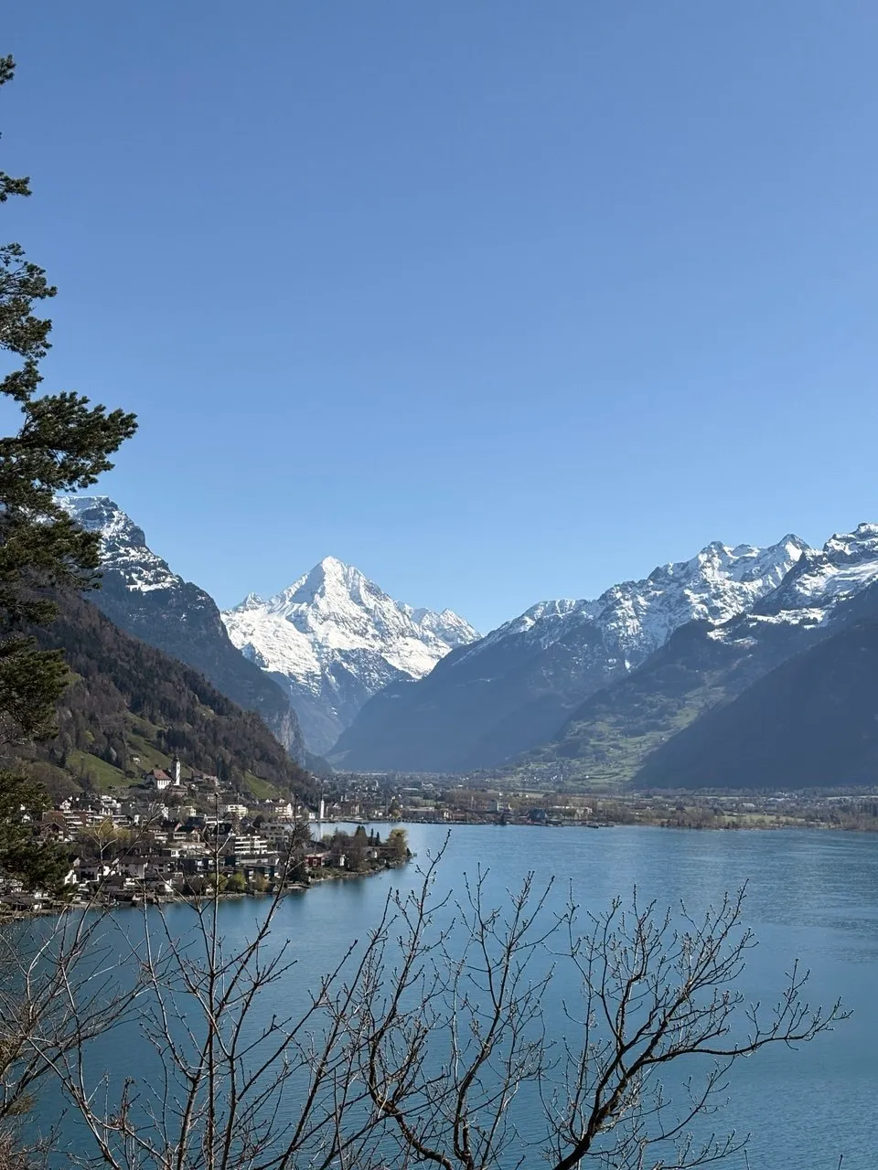 View across the blue lake to a snow-capped pyramid peak, with a village and church tower on the far shore and bare branches in the foreground.