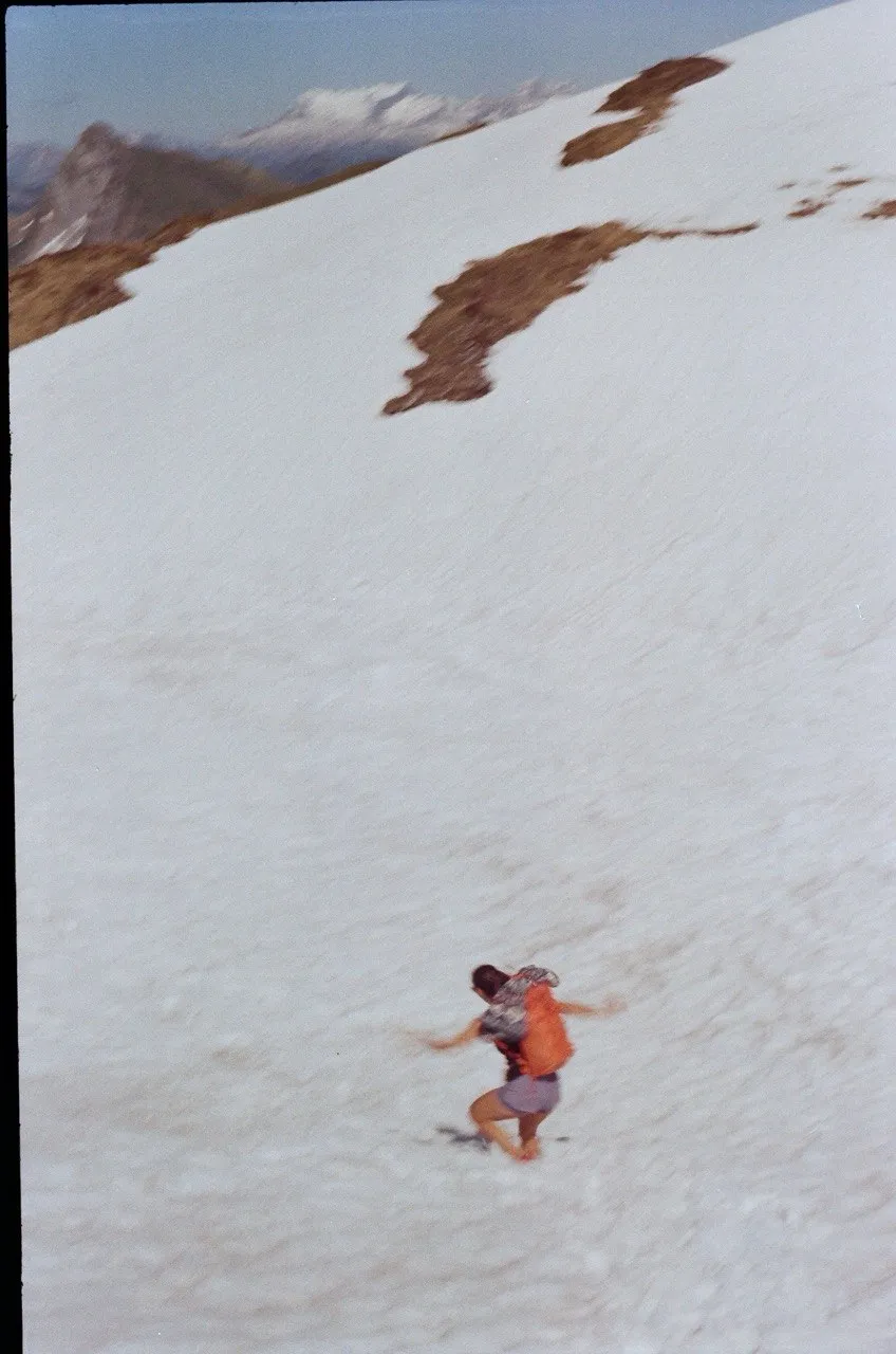 A hiker striding down a wide snowfield high on the ridge, with mountains in the hazy distance.