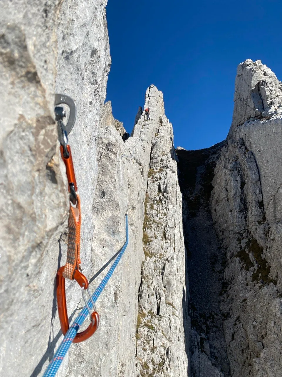 An orange quickdraw clipped to a rope on grey rock, jagged limestone towers and a tiny climber beyond under a deep blue sky.