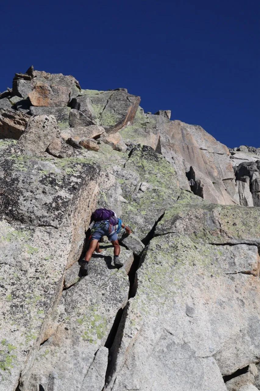 A scrambler in shorts climbs a steep light-grey granite slab beneath a deep blue sky.