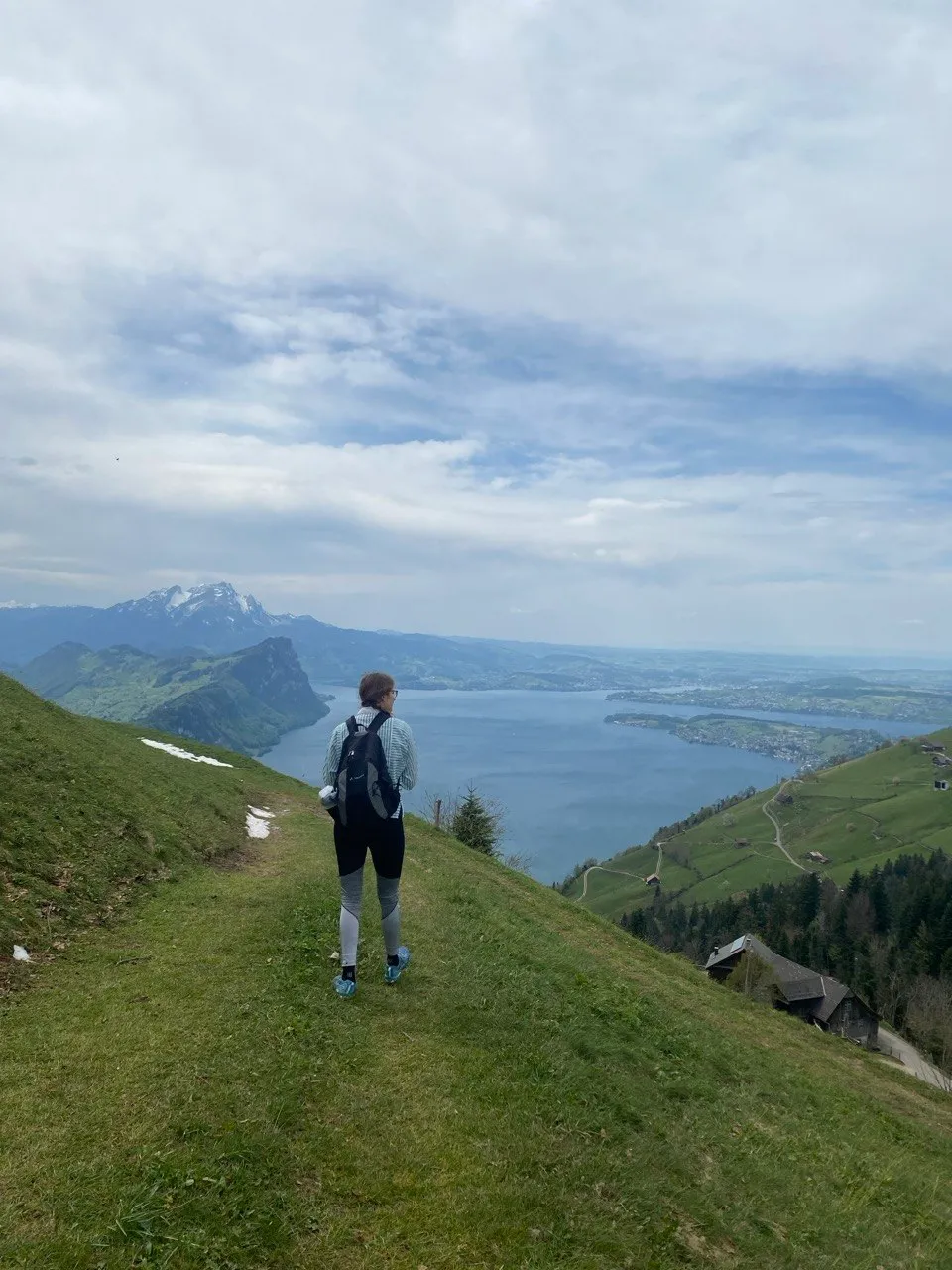 A hiker looks down from a grassy ridge onto the deep blue waters of Lake Lucerne, with snow-capped peaks and a farmhouse on the slope below.