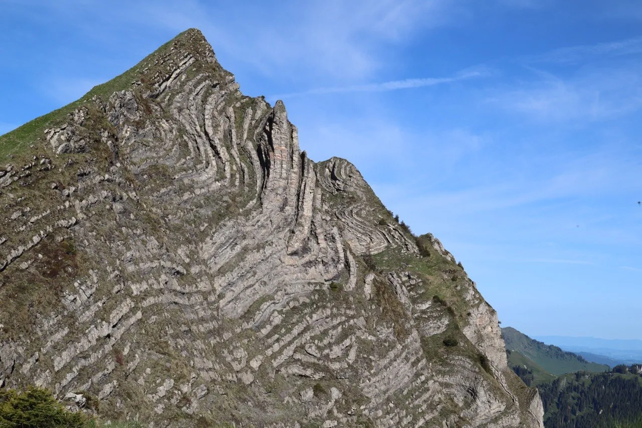A sharply folded rock peak with dramatic layered strata rises against a clear blue sky.