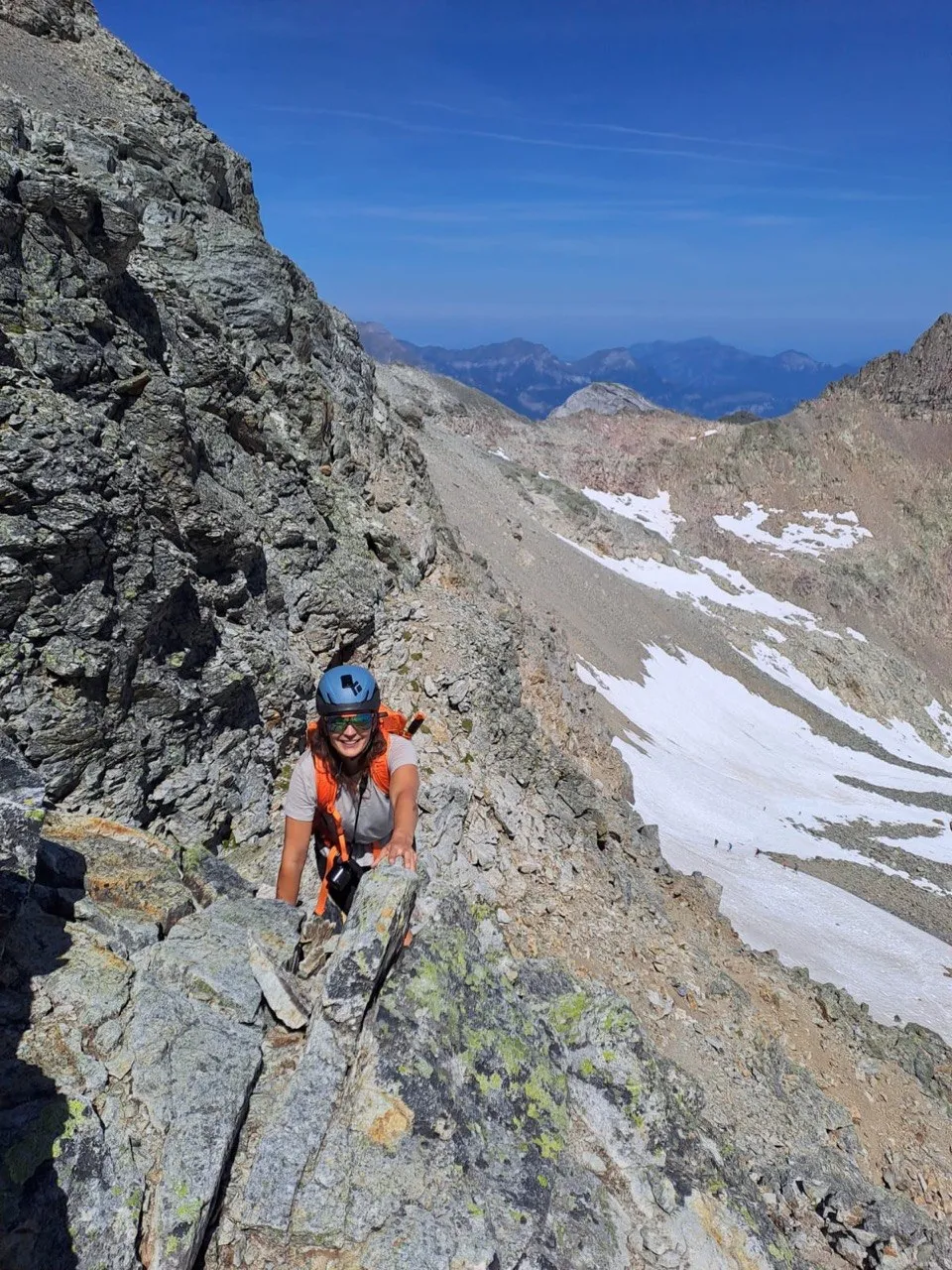 A smiling hiker climbs a rocky ridge, snowfields and distant peaks visible behind her.