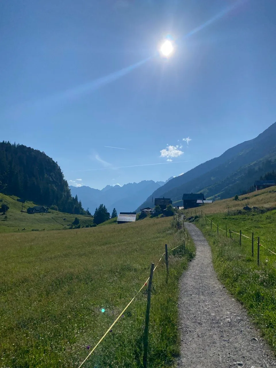 A sunlit mountain trail passes blooming meadows and rustic alpine huts, with rugged peaks in the backlight.