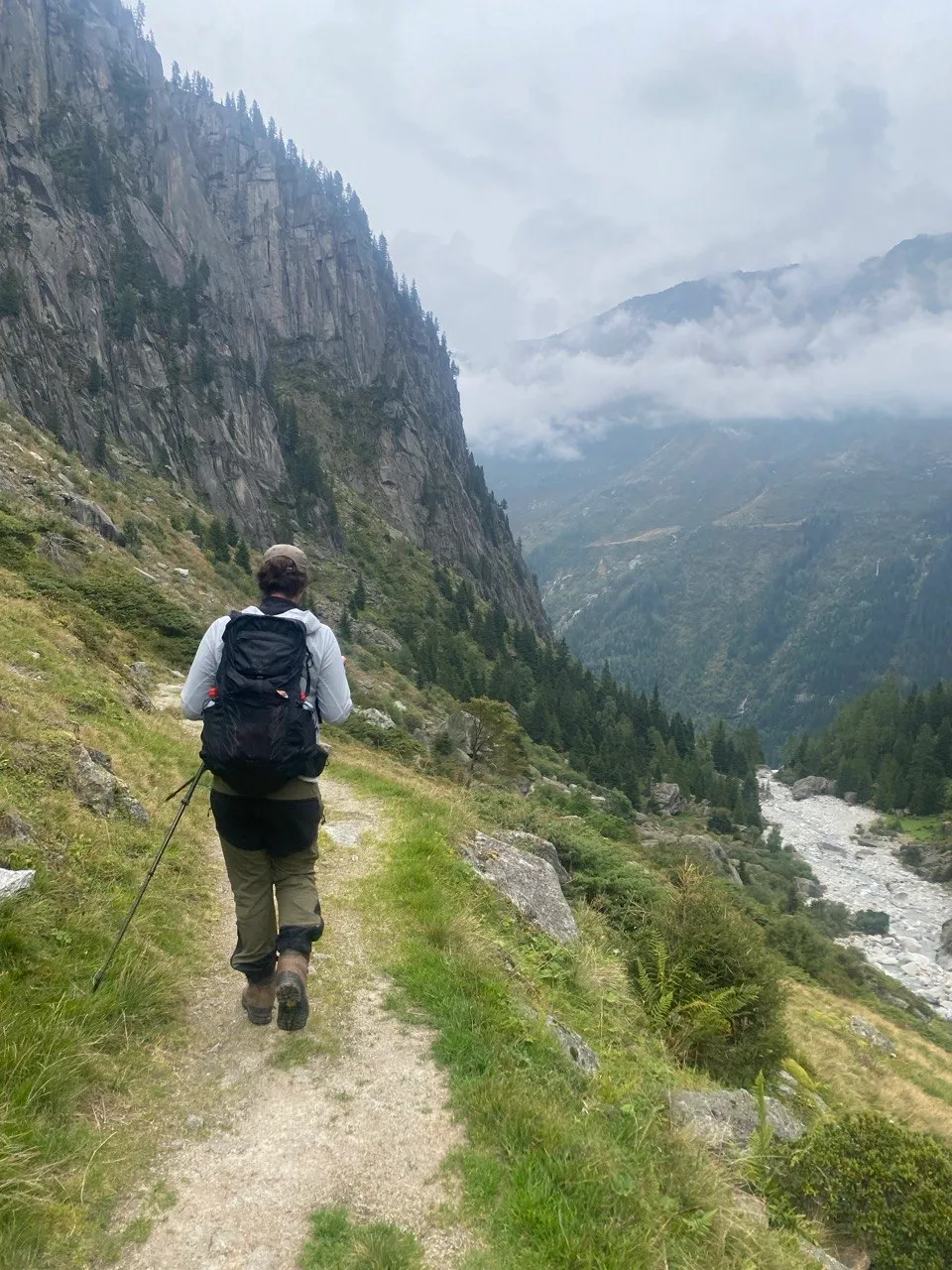 A hiker walks along a narrow trail on a steep mountain flank, with a river valley and fog-covered peaks below.