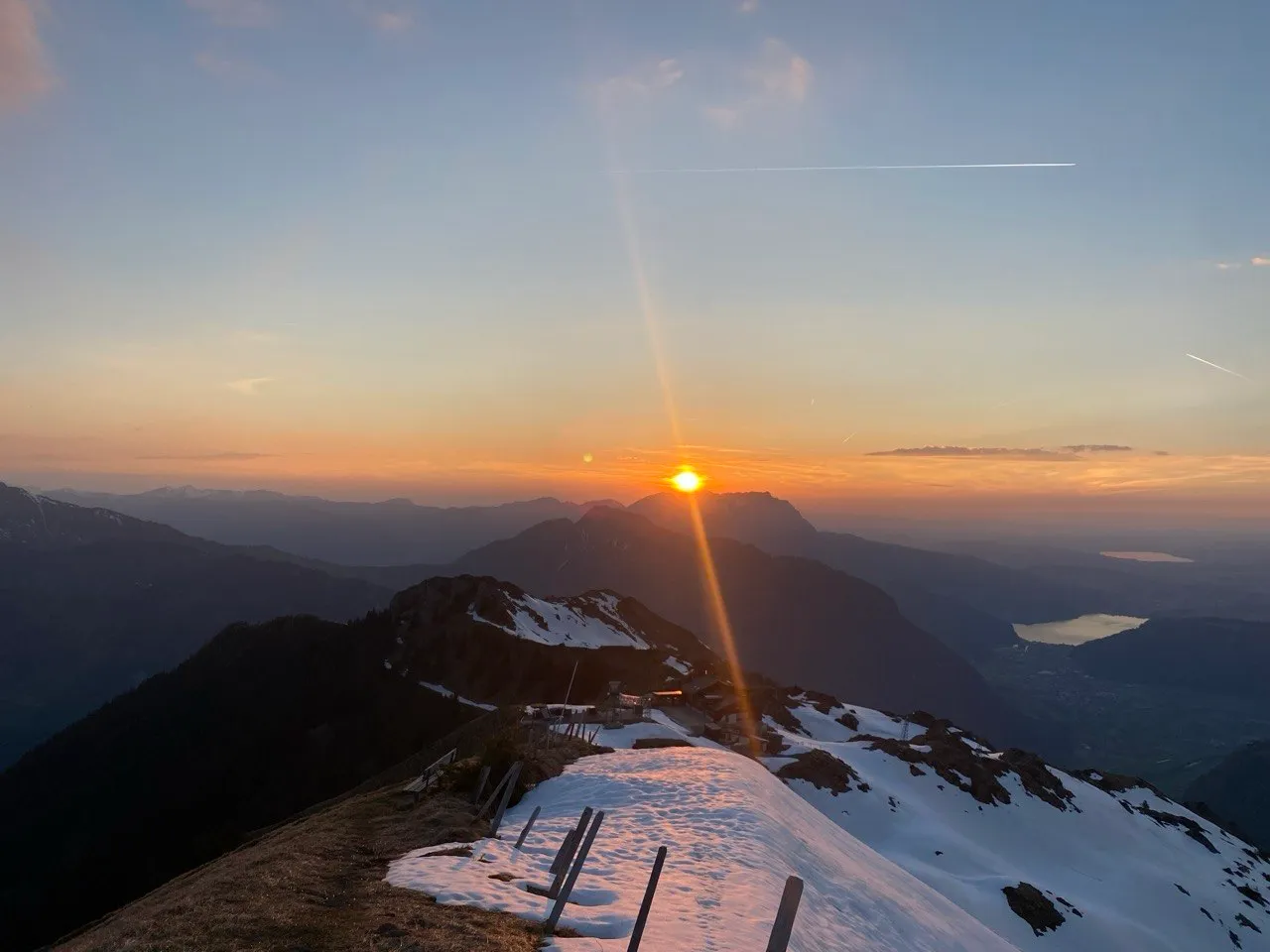 Sunset over the mountains, the sun just above a ridge, with lakes shimmering in the haze far below.