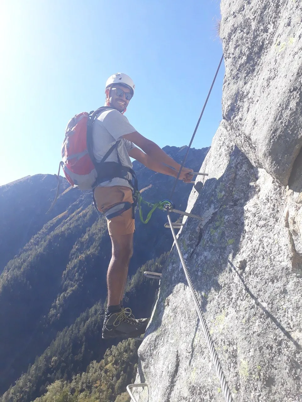 A climber with a red pack stands on metal rungs on a steep rock face, reaching for the steel cable, forested slopes far below.