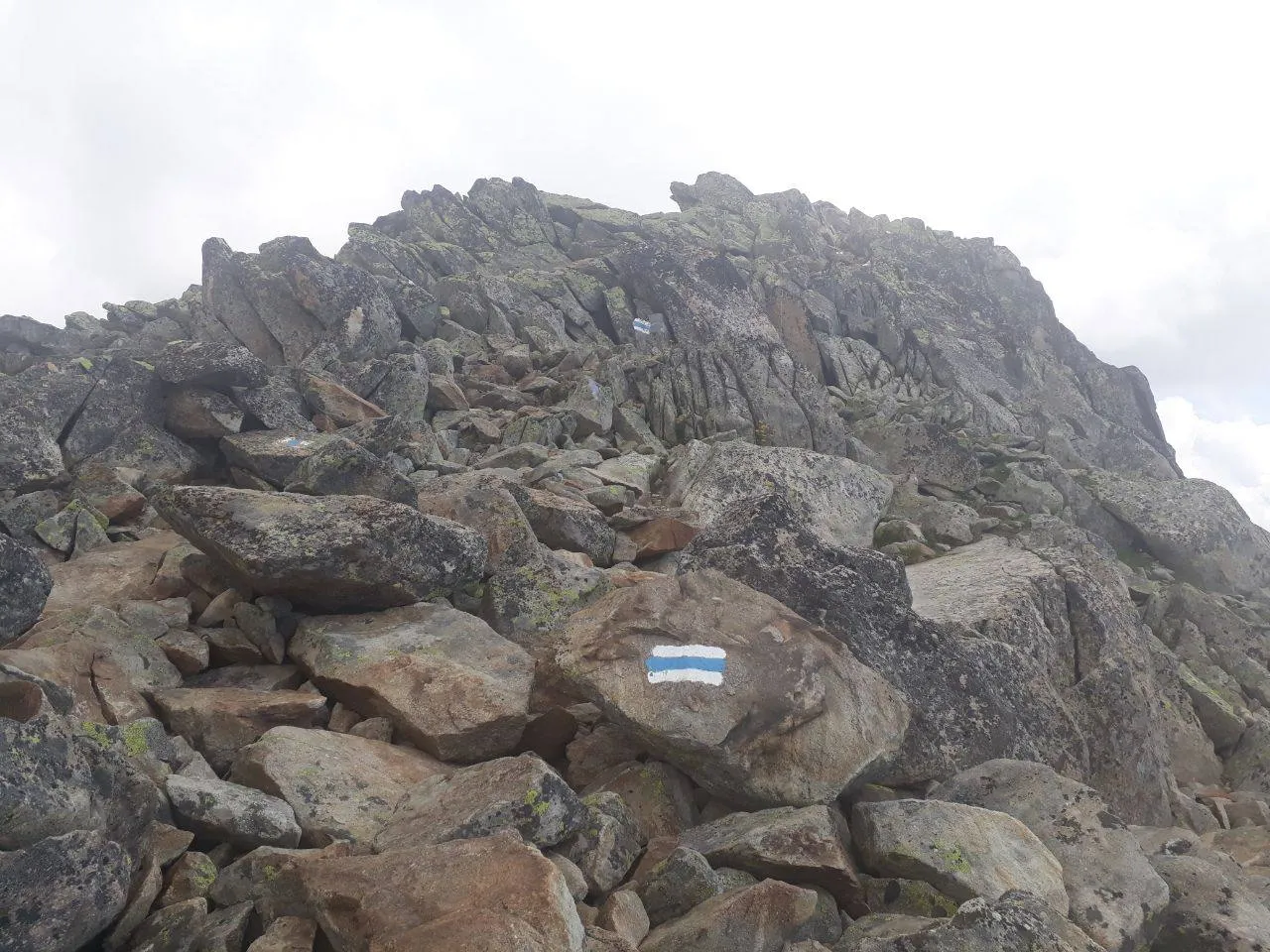 Coarse, lichen-covered granite blocks with blue-and-white trail markers, the jagged summit rising above under an overcast sky.