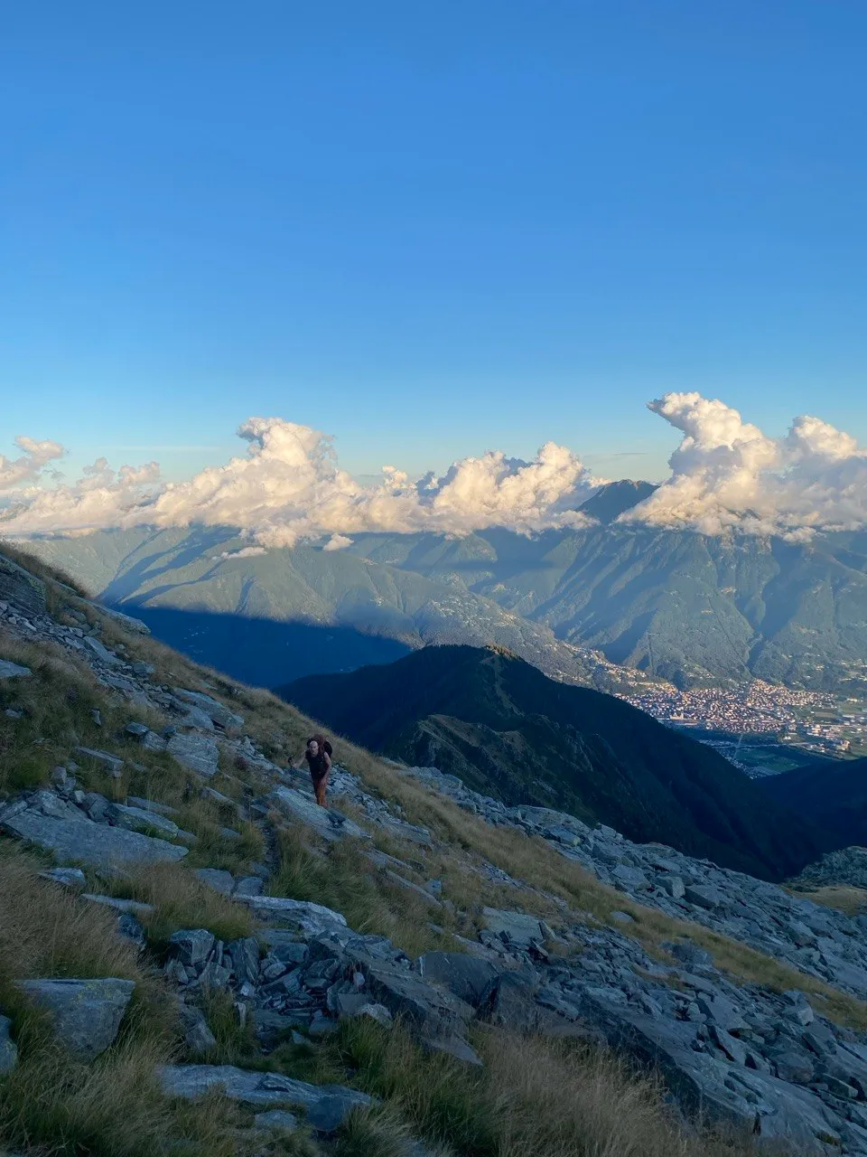 A hiker ascends a steep grassy ridge high above the valley, cloud-capped southern-Alpine peaks and the town of Bellinzona in the distance.