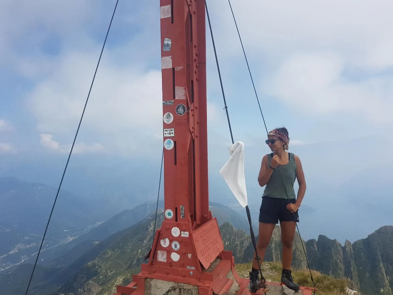 A hiker stands at the red summit cross of the Gridone, with hazy mountain ranges and Lake Maggiore far below under a cloudy sky.