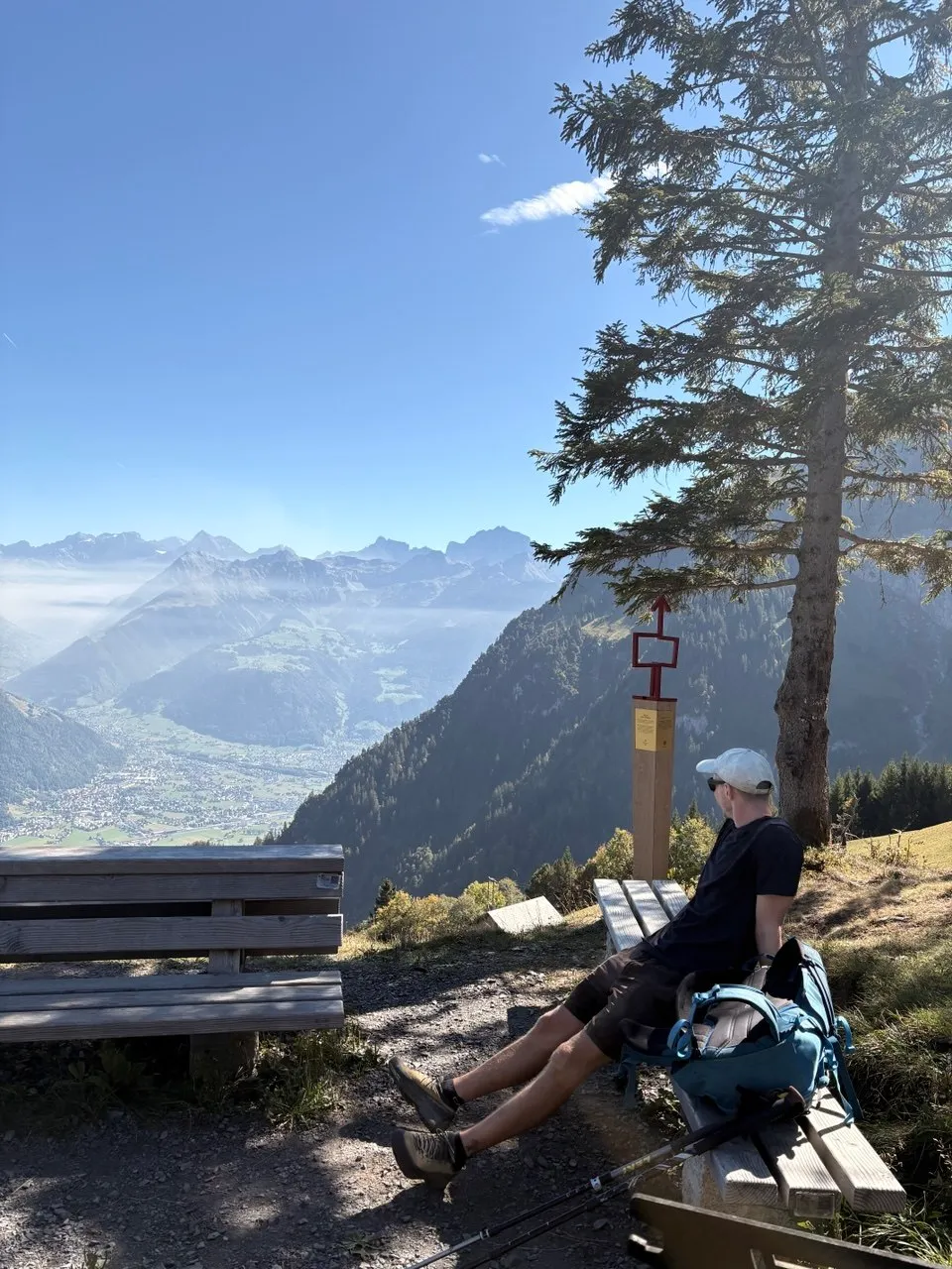 A hiker rests on a wooden bench at the barbecue spot with a wide view over the Reuss valley and the Uri Alps.