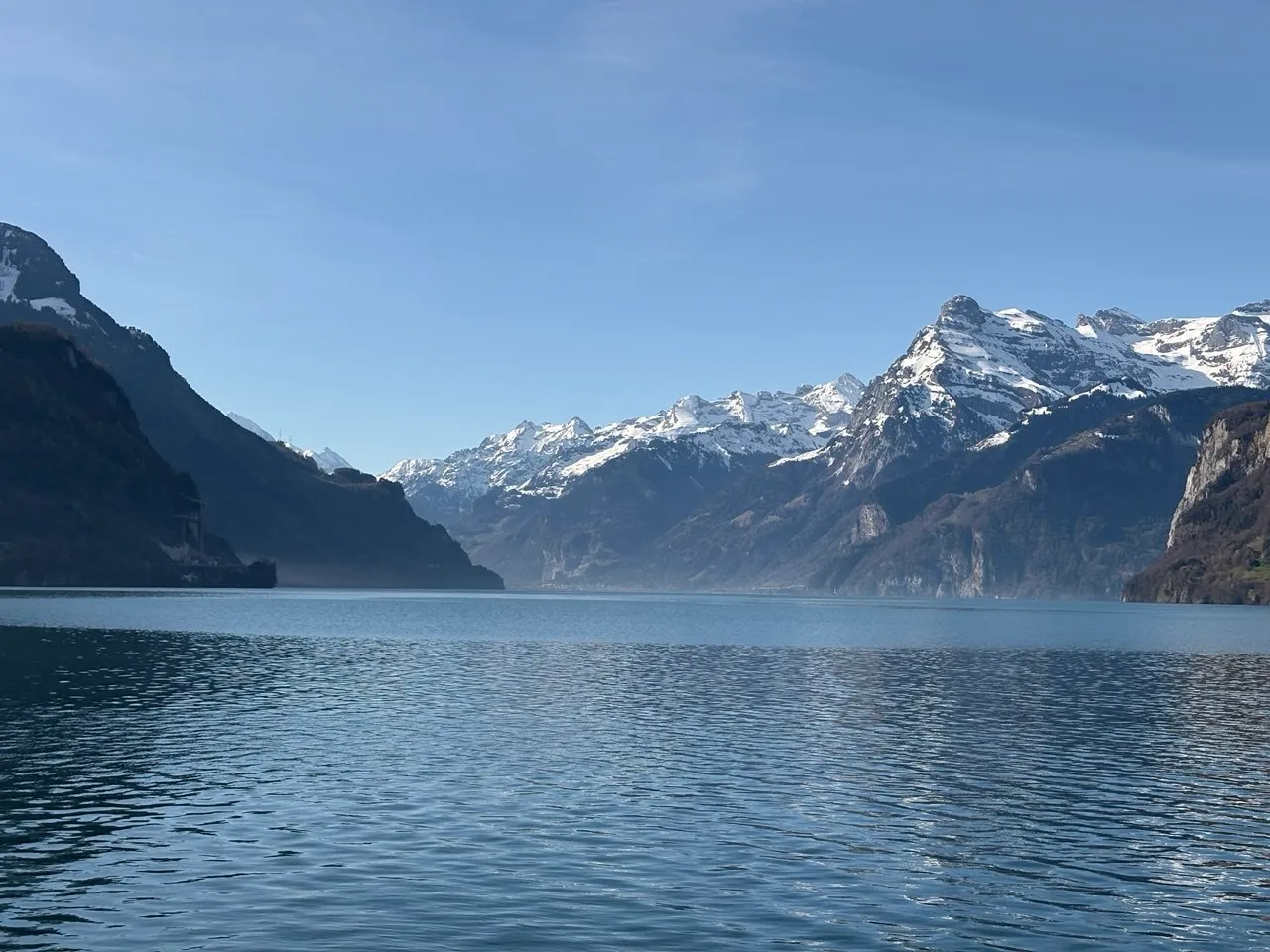 A wide view from lake level across Lake Lucerne to a long chain of snow-capped mountains under a blue sky.