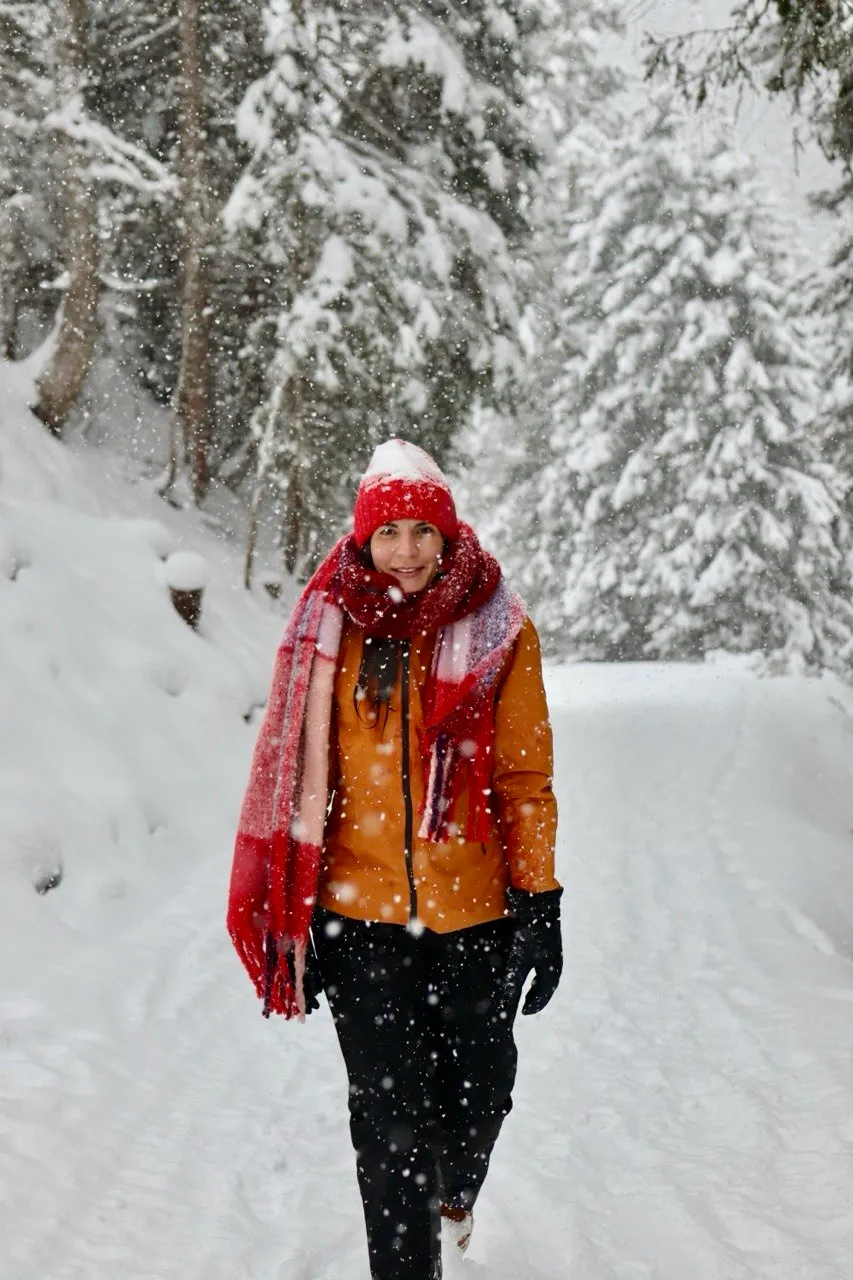 A smiling hiker in a mustard jacket with a red knit hat and scarf walks a snowy forest path through falling snow.