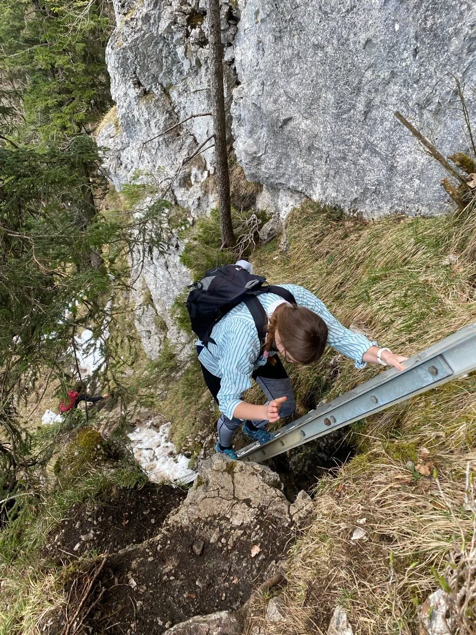 A hiker climbs a small metal ladder across a steep, grassy passage beneath a rock face.