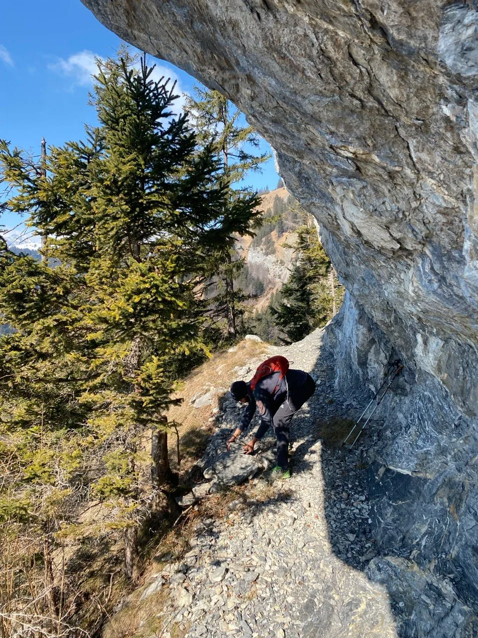 A hiker scrambles over scree beneath an overhanging rock roof, surrounded by larches with distant peaks in view.