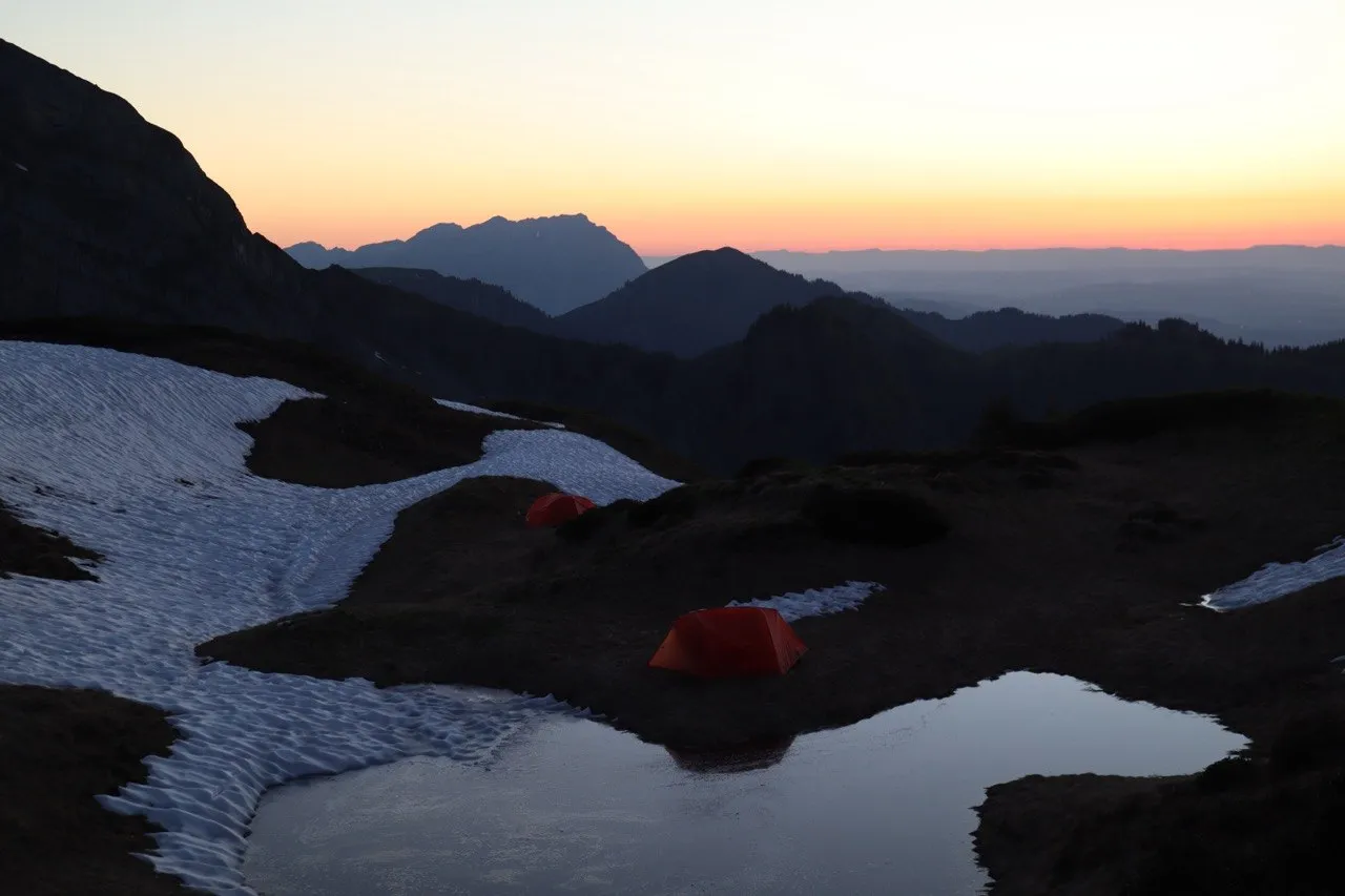 Two small tents pitched among snow patches on the ridge, glowing orange at dusk with mountains fading into the distance.