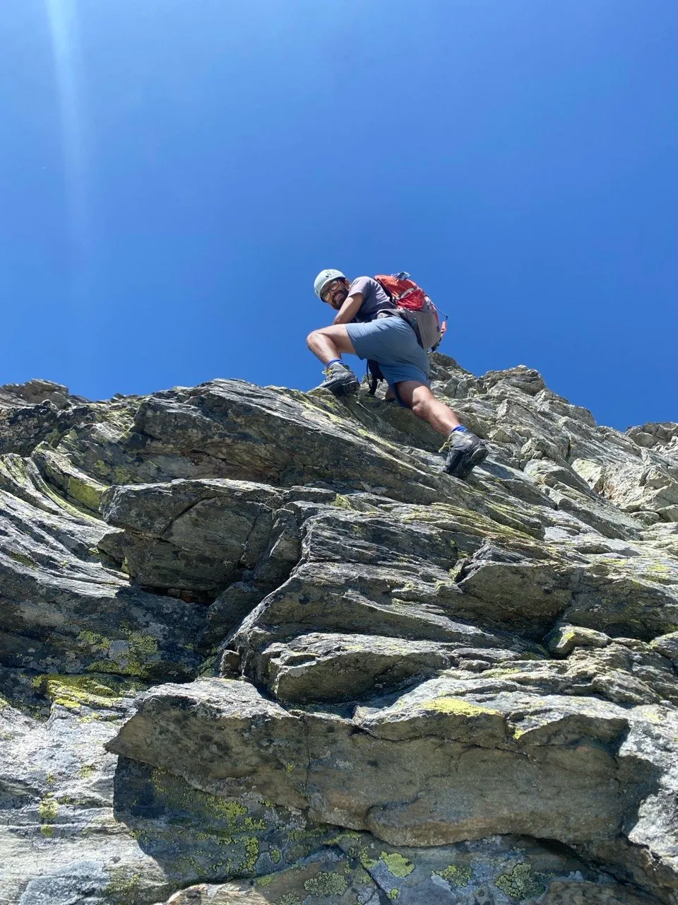 Shot from below, a hiker climbs over jagged rock slabs against a bright blue sky.