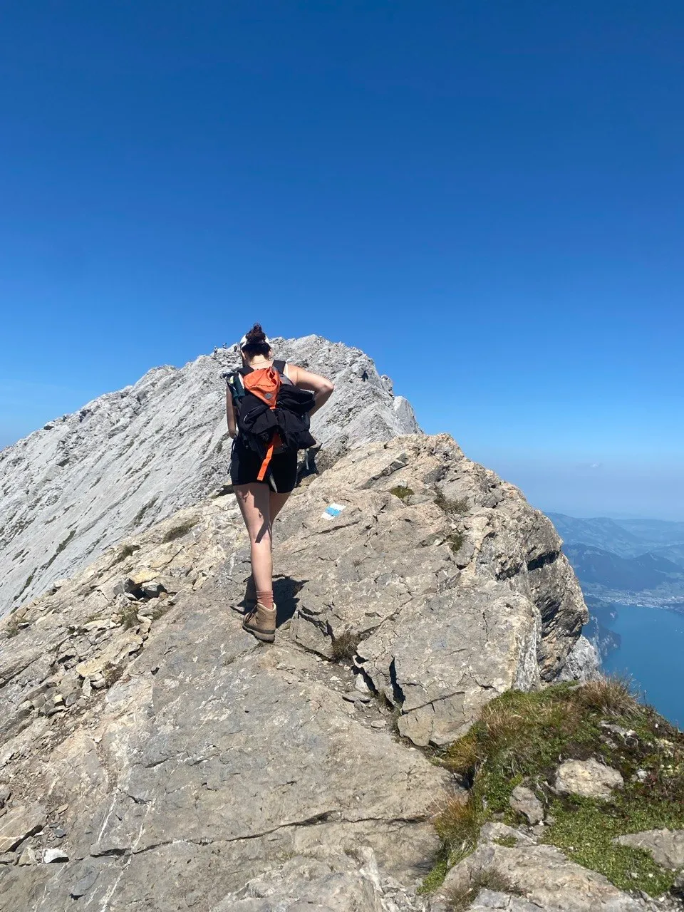 A hiker stands on a narrow, exposed rock ridge high above turquoise Lake Uri.