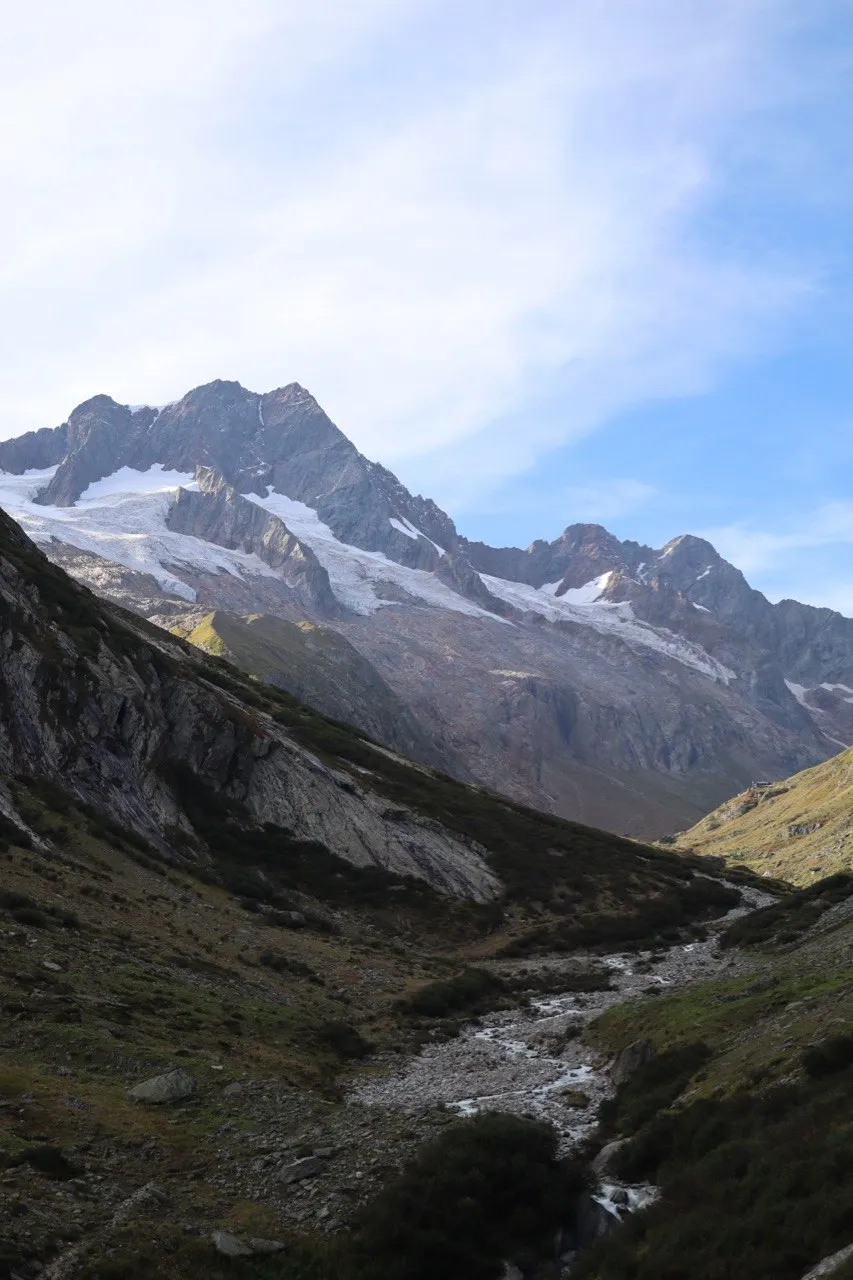 A snow-streaked mountain peak rises above a rocky valley with a stream, under a blue sky.