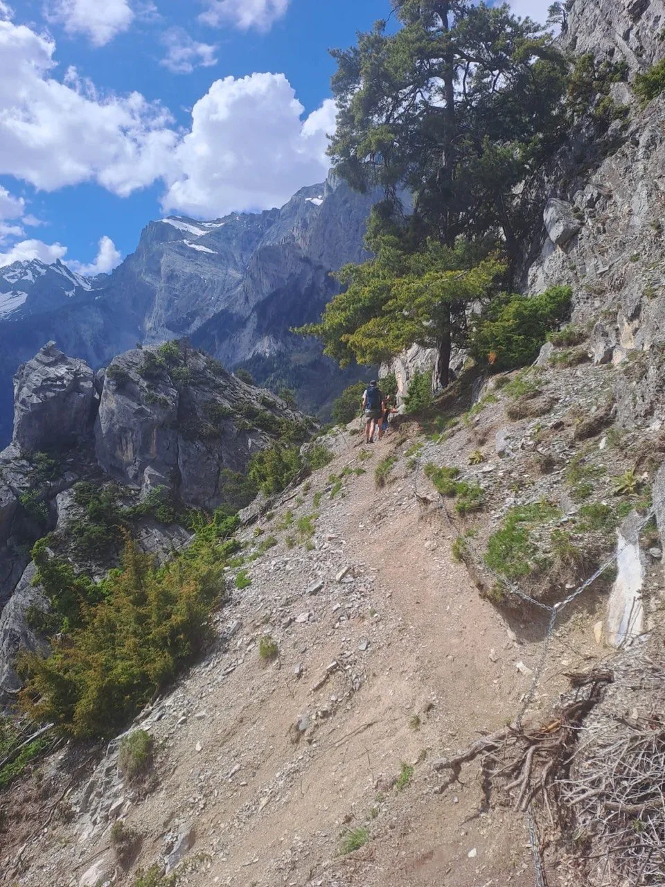 A hiker on a narrow ridge trail with jagged, partly snow-streaked limestone peaks rising in the background.