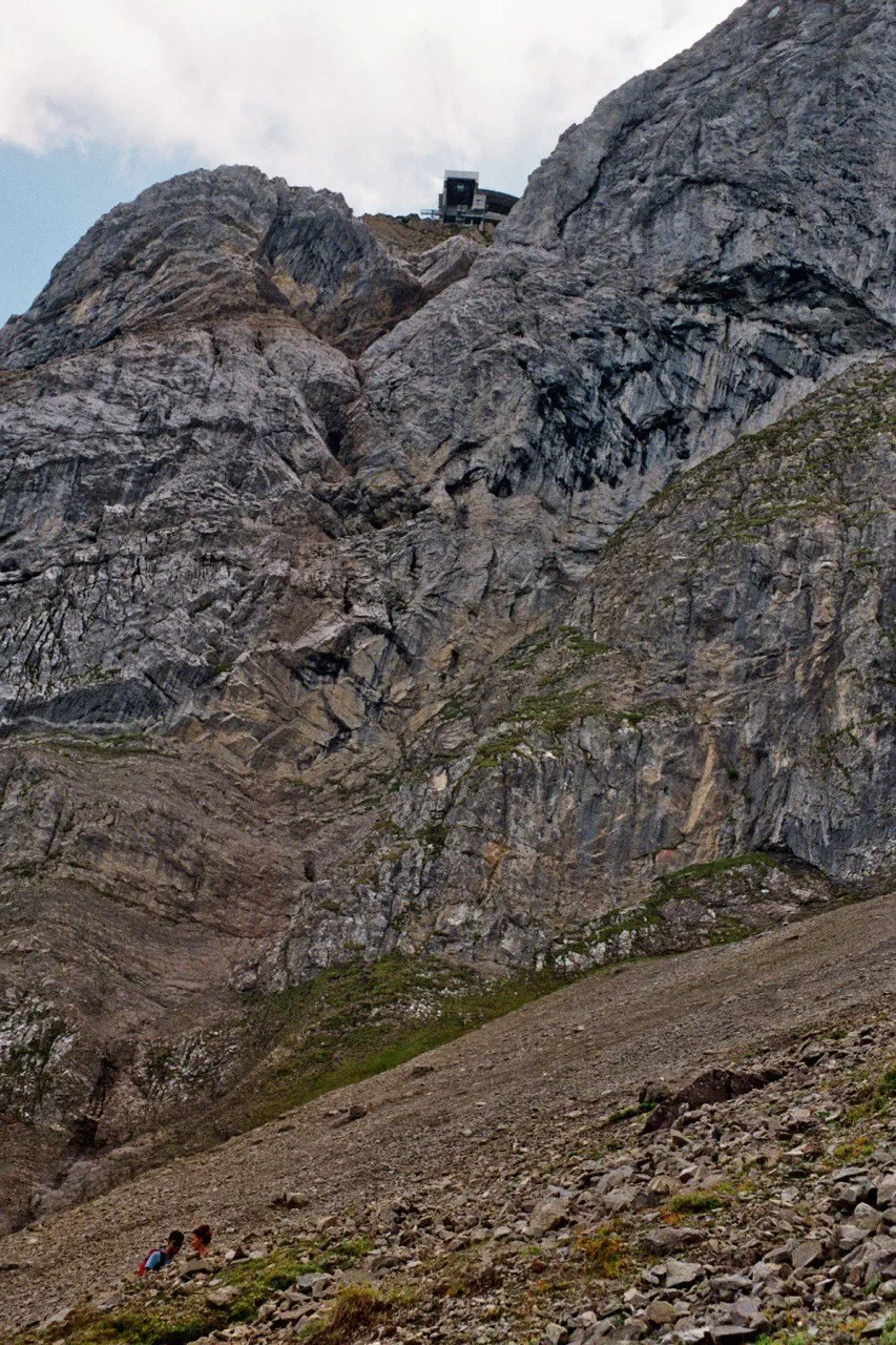 The summit station perched on a rock rib high above a vast face of scree and cliff, two tiny hikers below.